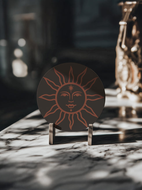 Round ceramic coaster in deep black featuring an intricate burnt-orange sun illustration with a serene, symmetrical face, radiating curved rays, and a delicate star detail on the forehead. The smooth, matte surface enhances the contrast between the dark background and the warm-toned design, creating a celestial, vintage-inspired aesthetic.